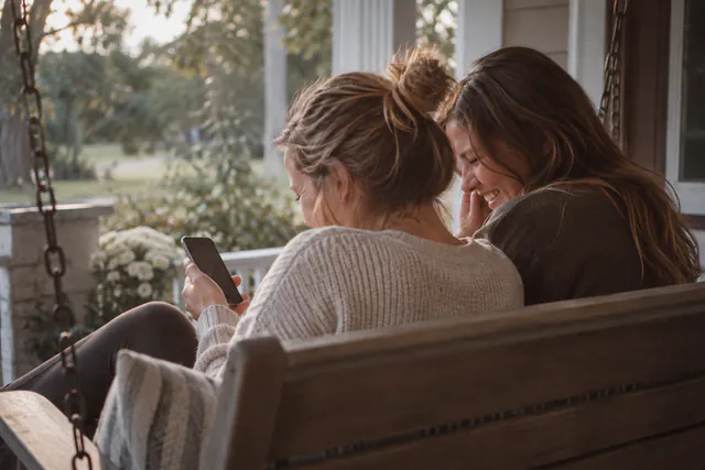 AI-generated photo: two women sit close on a porch swing, seen from behind, one holding a phone while the other laughs.