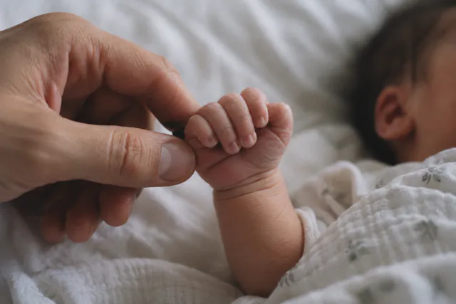 AI-generated photo: a newborn’s hand holds a parent’s fingertip, the baby’s face soft in the background.